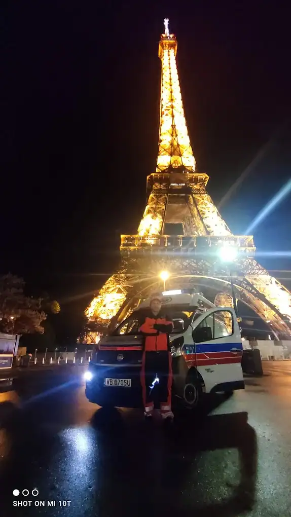 Międzynarodowy Transport Medyczny Włocławek – Cała Europa w Zasięgu Ręki Propozycja: Ratownik medyczny stoi przed ambulansem z otwartymi drzwiami, na tle oświetlonej Wieży Eiffla w Paryżu. Międzynarodowy transport medyczny Włocławek.