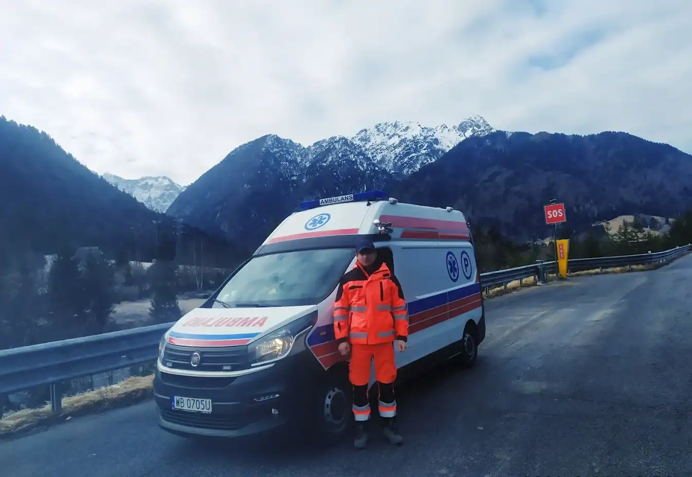 Ambulans na górskiej drodze z ratownikiem w stroju odblaskowym, widok na wysokie, śnieżne góry w tle, zapewniający transport osób niepełnosprawnych w trudnych warunkach terenowych.
